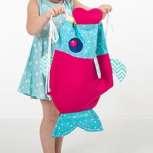 May include: A colourful, fish-shaped drawstring bag. The bag has a bright pink body, a blue polka-dot top, and a blue tail fin. The fish has a red button eye and a heart-shaped detail. White drawstrings are visible.