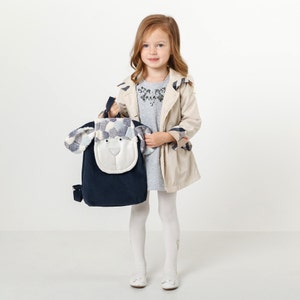 Könnte beinhalten: Ein junges Mädchen trägt einen beigefarbenen Trenchcoat mit blau-weißem Karomuster, ein graues Kleid und weiße Strumpfhosen. Sie hält einen blauen Rucksack mit einem weißen und grauen Hundegesicht auf der Vorderseite.
