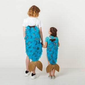 May include: Two children wearing blue and brown fishtail costumes stand back to back with their hands clasped. The costumes are made of fabric and have a scaly texture.