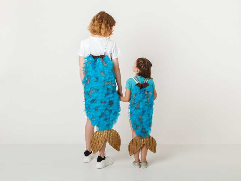 May include: Two children wearing blue and brown fishtail costumes stand back to back with their hands clasped. The costumes are made of fabric and have a scaly texture.