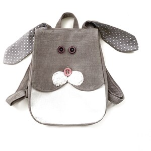 May include: A small grey linen backpack with bunny ears. The ears are lined with white polka dots on a grey background. The backpack has a white front pocket and a pink button nose with white felt whiskers. The backpack has two brown buttons for eyes.