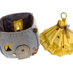 May include: A gray felt house with a yellow felt roof and a yellow felt door. The house has a yellow felt window with curtains. The house is sitting on a white surface. There is a yellow felt dress with a brown ribbon and ladybug buttons on the dress.
