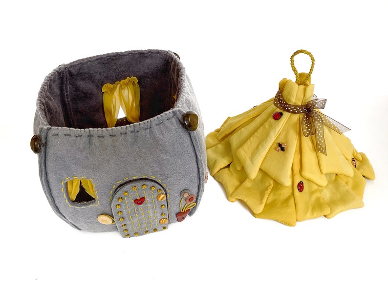 May include: A gray felt house with a yellow felt roof and a yellow felt door. The house has a yellow felt window with curtains. The house is sitting on a white surface. There is a yellow felt dress with a brown ribbon and ladybug buttons on the dress.