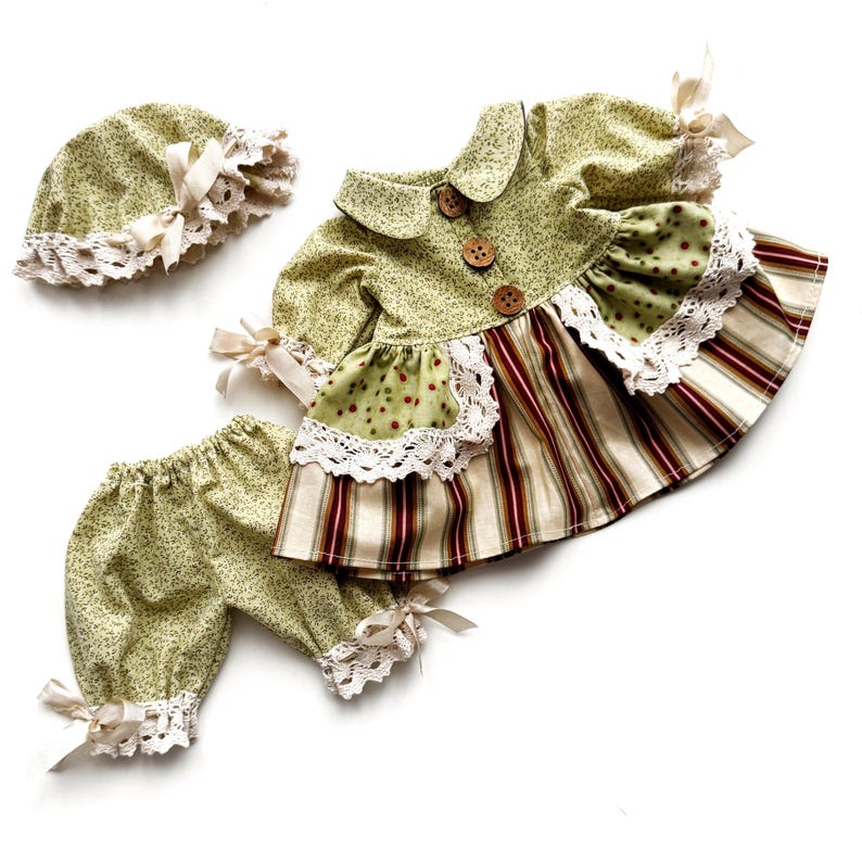 May include: A green and white floral print baby dress with a white lace trim and brown buttons. The dress is paired with matching green and white floral print bloomers and a green and white floral print hat with a white lace trim.
