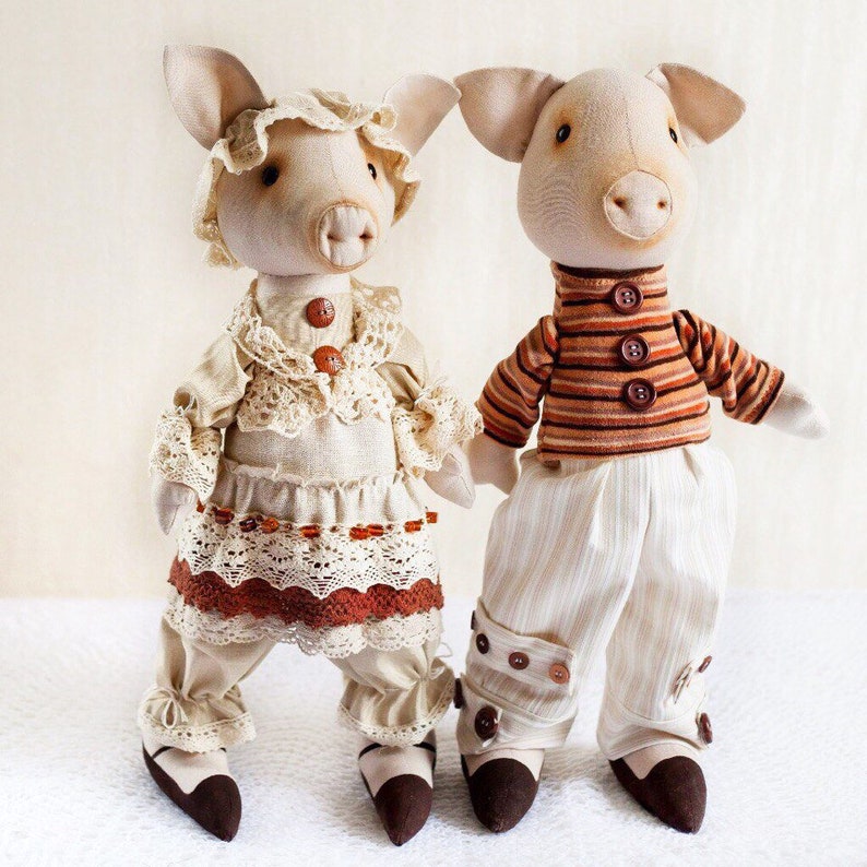 May include: Two handmade fabric pig dolls. The female pig doll is wearing a white dress with brown lace trim and brown shoes. The male pig doll is wearing a striped shirt, white pants with brown buttons, and brown shoes.