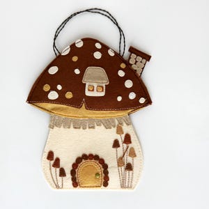 May include: A whimsical felt mushroom house with a brown cap dotted with white circles. The house features a cream-colored body, a small door, and a chimney. A black string is attached for hanging.