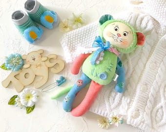 May include: A green and pink patchwork toy with a cat face and blue bow. The toy is lying on a white knitted blanket with a wooden sign that says "Toys".