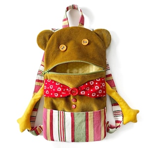 May include: A brown plush backpack shaped like a bear with a red bow tie and a zippered opening. The backpack has a striped fabric design with red, white, and green stripes.