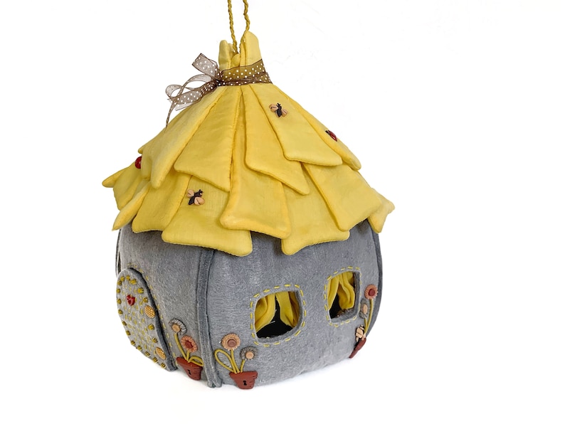 May include: A gray felt house with a yellow felt roof and a small door. The roof is decorated with small yellow bees and red ladybugs. The house has two windows with yellow curtains. The house is decorated with small yellow flowers and brown buttons.