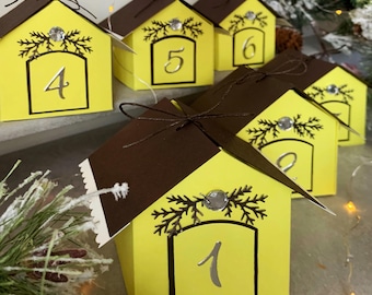 May include: Yellow house-shaped boxes with brown roofs, numbered 1 to 6. Each box features silver accents, a sprig design, and a small bow. The boxes are designed to resemble miniature houses.