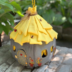 May include: A gray felt fairy house with a yellow felt roof, decorated with embroidered flowers and ladybugs. The roof is gathered and tied with a brown ribbon.