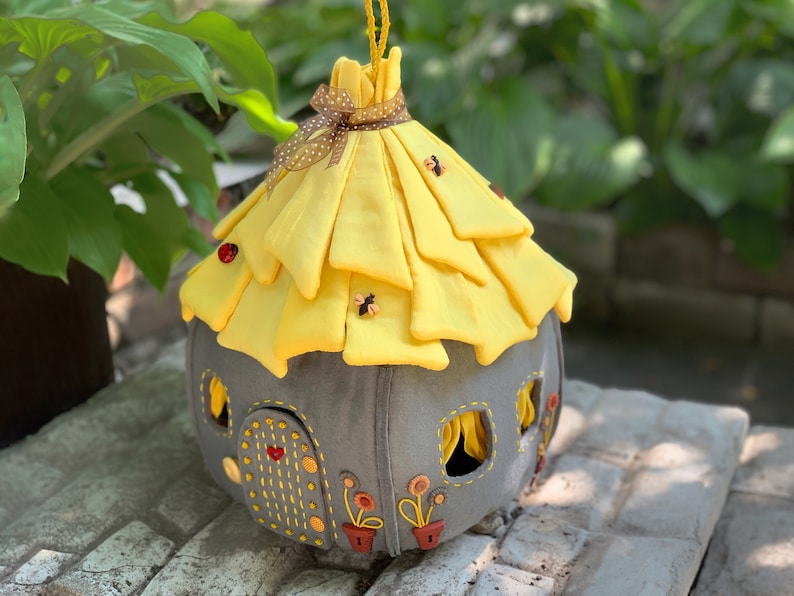 May include: A gray felt fairy house with a yellow felt roof, decorated with embroidered flowers and ladybugs. The roof is gathered and tied with a brown ribbon.