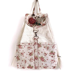 May include: A white quilted backpack with a floral print on the bottom half. The backpack has a flap with a decorative floral embellishment in shades of red, brown, and beige.