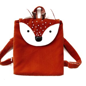 May include: A small, orange, plush backpack with a white fox face and antlers. The backpack has white polka dots on the front.