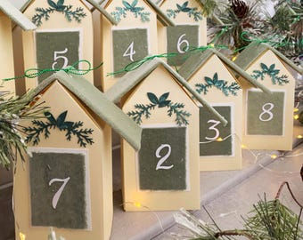 May include: A series of miniature house-shaped boxes, each numbered. The boxes are cream-coloured with green roofs and green squares displaying white numbers. They are decorated with green leaf and branch accents, arranged in a row.