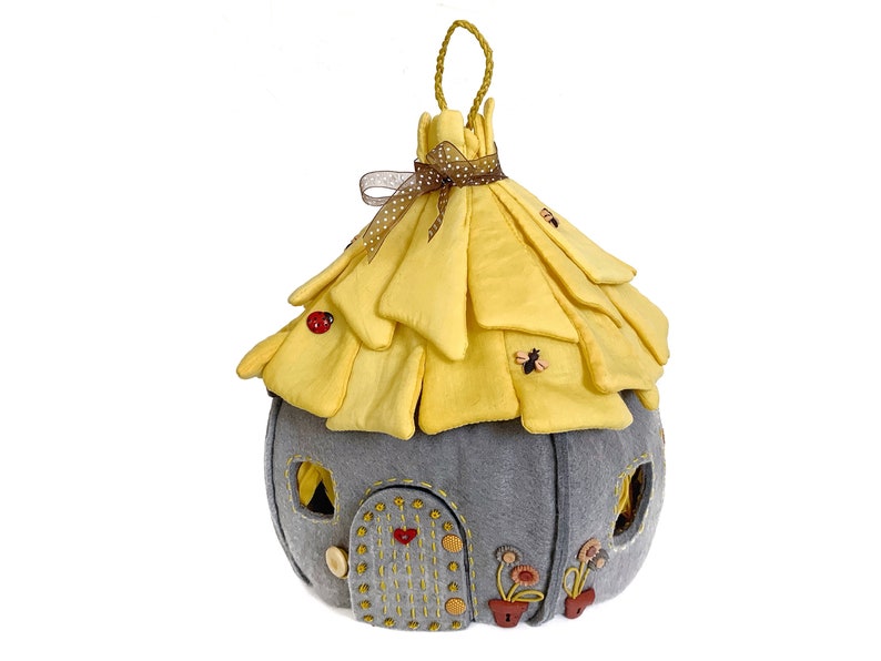 May include: A gray felt fairy house with a yellow felt roof, decorated with embroidered details and fabric flowers. The roof has a brown ribbon and a loop for hanging.