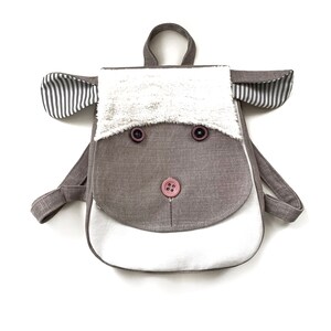 May include: A brown and white linen backpack with a sheep face design. The backpack has two straps and a white fur trim around the face. The sheep face has two brown buttons for eyes and a pink button for a nose.