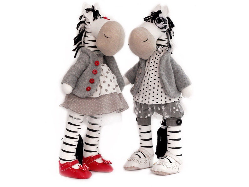May include: Two plush zebra toys wearing dresses and jackets. One zebra has red shoes and the other has white shoes.