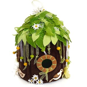 K&ouml;nnte beinhalten: Ein braunes Filz-Vogelhaus mit einem gr&uuml;nen Filzdach, verziert mit wei&szlig;en Filzblumen, gelben und gr&uuml;nen Perlen sowie wei&szlig;en und braunen Filzv&ouml;geln. Das Vogelhaus hat eine runde &Ouml;ffnung mit einem braunen Filzrahmen.