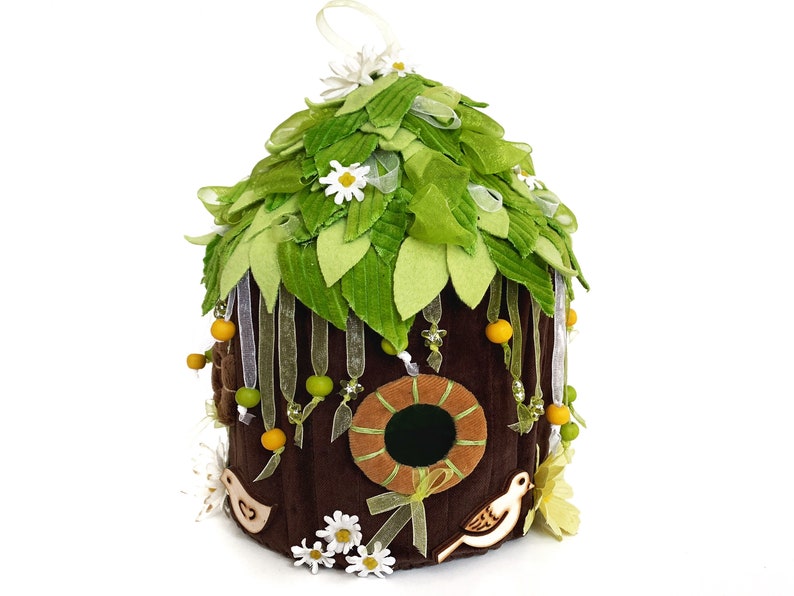 K&ouml;nnte beinhalten: Ein braunes Filz-Vogelhaus mit einem gr&uuml;nen Filzdach, verziert mit wei&szlig;en Filzblumen, gelben und gr&uuml;nen Perlen sowie wei&szlig;en und braunen Filzv&ouml;geln. Das Vogelhaus hat eine runde &Ouml;ffnung mit einem braunen Filzrahmen.