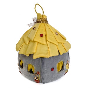 May include: A yellow felt fairy house with a gray felt base. The house has a pointed roof with a brown ribbon and white polka dots. The house has several windows and is decorated with red and brown buttons.