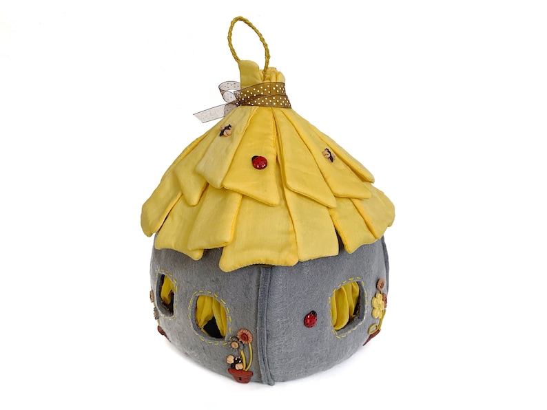 May include: A yellow felt fairy house with a gray felt base. The house has a pointed roof with a brown ribbon and white polka dots. The house has several windows and is decorated with red and brown buttons.