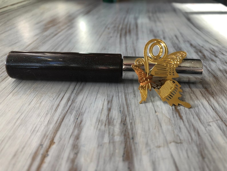 May include: A gold butterfly bookmark with a decorative swirl design. The bookmark is attached to a black and silver pen.