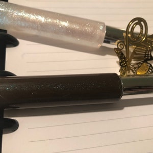 May include: Two tubes of lip gloss, one with a clear iridescent finish and the other with a dark brown finish. Both tubes have a silver metal cap.