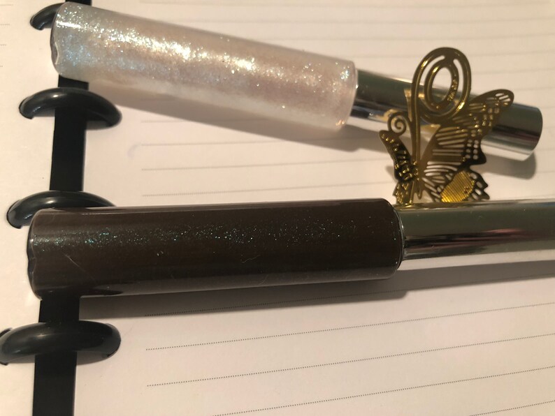 May include: Two tubes of lip gloss, one with a clear iridescent finish and the other with a dark brown finish. Both tubes have a silver metal cap.
