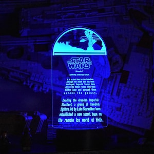 Puede incluir: Un letrero iluminado azul con el texto "STAR WARS EMPIRE STRIKES BACK Episodio V" y una descripción de la trama de la película. El letrero tiene forma de media luna con un planeta en el fondo.