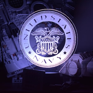 May include: A white acrylic light-up sign with the United States Navy seal. The seal features an eagle with outstretched wings, a shield with stars, and an anchor. The words "United States Navy" are written around the seal.