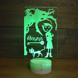 Op de afbeelding: Een groen LED-nachtlampje met een wit acryl paneel met een silhouet van een meisje, een kat, een sleutel en een knoop. De naam van het meisje, "Alayna", staat boven de sleutel.