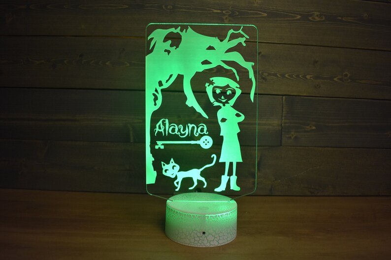 Op de afbeelding: Een groen LED-nachtlampje met een wit acryl paneel met een silhouet van een meisje, een kat, een sleutel en een knoop. De naam van het meisje, "Alayna", staat boven de sleutel.