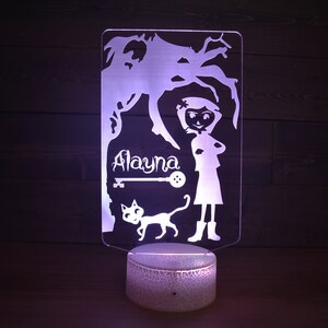 Op de afbeelding: Een wit acryl nachtlampje met een silhouet van een meisje, een kat, een sleutel en de naam "Alayna". Het lampje staat op een witte basis met een getextureerd oppervlak.