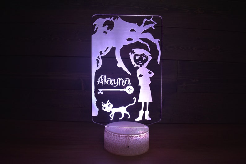Op de afbeelding: Een wit acryl nachtlampje met een silhouet van een meisje, een kat, een sleutel en de naam "Alayna". Het lampje staat op een witte basis met een getextureerd oppervlak.