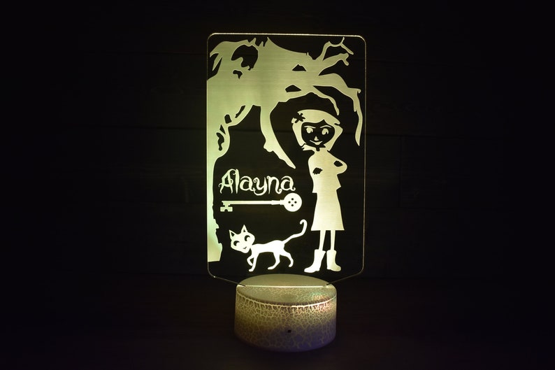 Op de afbeelding: Een wit acryl nachtlampje met een silhouet van een meisje, een kat, een sleutel en de naam "Alayna". Het lampje staat op een ronde, getextureerde basis.
