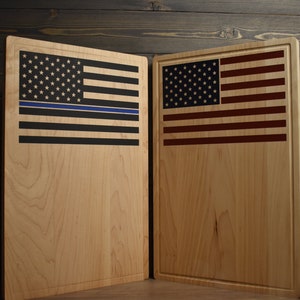 May include: Two wooden cutting boards with American flag designs. The board on the left has a black and blue American flag with a thin blue line, while the board on the right has a red, white, and blue American flag.