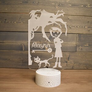 Op de afbeelding: Een wit acryl nachtlampje met een silhouet van een meisje, een kat en een boom. Het meisje draagt een jurk en laarzen. De naam "Alayna" staat op het lampje.