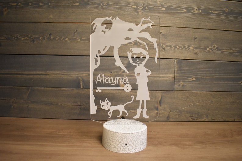 Op de afbeelding: Een wit acryl nachtlampje met een silhouet van een meisje, een kat en een boom. Het meisje draagt een jurk en laarzen. De naam "Alayna" staat op het lampje.