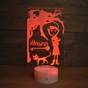 Op de afbeelding: Een rood oplichtend nachtlampje met een witte basis. Het lampje heeft een silhouet van een meisje met een kat en een boom. De naam "Alayna" staat op het lampje.