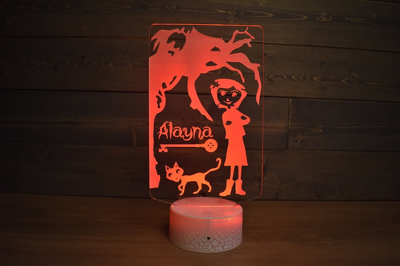 Op de afbeelding: Een rood oplichtend nachtlampje met een witte basis. Het lampje heeft een silhouet van een meisje met een kat en een boom. De naam "Alayna" staat op het lampje.