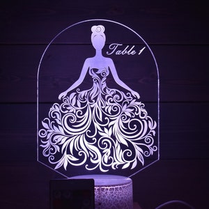 Op de afbeelding: Een wit silhouet van een vrouw in een jurk met bloemenmotieven, verlicht van achteren. Het silhouet bevindt zich in een helder acrylframe met de tekst "Tafel 1" bovenaan. De voet van de lamp is een getextureerde witte cilinder.