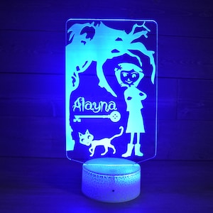 Op de afbeelding: Een blauw licht verlicht een wit acryl nachtlampje met een silhouet van een meisje, een kat, een sleutel en de naam "Alayna".