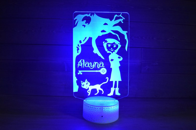 Op de afbeelding: Een blauw licht verlicht een wit acryl nachtlampje met een silhouet van een meisje, een kat, een sleutel en de naam "Alayna".