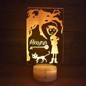 Op de afbeelding: Een geel nachtlampje met een silhouet van een meisje, een kat en een boom. Het meisje staat met haar armen gekruist en de naam "Alayna" staat op het lampje.
