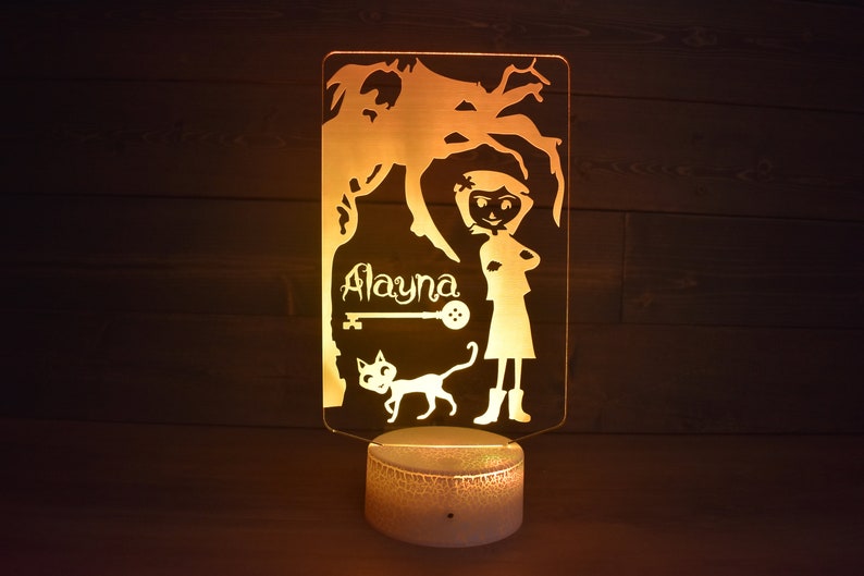 Op de afbeelding: Een geel nachtlampje met een silhouet van een meisje, een kat en een boom. Het meisje staat met haar armen gekruist en de naam "Alayna" staat op het lampje.