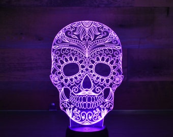 Könnte beinhalten: Ein violettes Licht beleuchtet einen weißen, kunstvoll gestalteten Zucker Schädel. Der Schädel ist aus Acryl und hat ein detailliertes Blumenmuster.