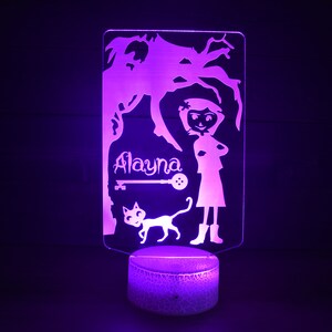 Op de afbeelding: Een paarse nachtlamp met een silhouet van een meisje, een kat en een boom. Het meisje staat met haar armen gekruist en de naam "Alayna" staat boven haar. Een sleutel en een knoop zijn ook in het ontwerp opgenomen.