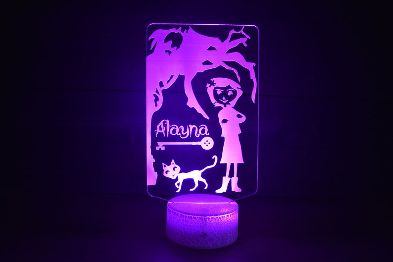 Op de afbeelding: Een paarse nachtlamp met een silhouet van een meisje, een kat en een boom. Het meisje staat met haar armen gekruist en de naam "Alayna" staat boven haar. Een sleutel en een knoop zijn ook in het ontwerp opgenomen.