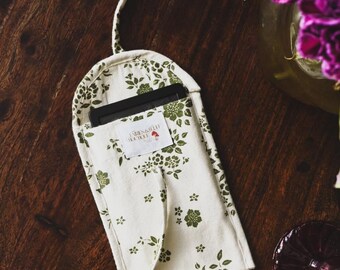Op de afbeelding: Een crèmekleurige wijnfleszak met een groen bloemenpatroon. De tas heeft een lange, dunne handgreep en een klein label met tekst. De tas ligt op een donker houten oppervlak, met een wijnglas en bloemen op de achtergrond.